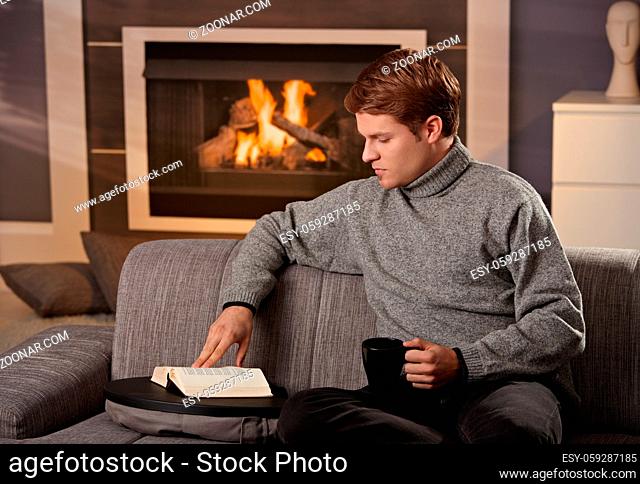 Young man sitting on sofa at home on a cold winter day, reading book in front of fireplace