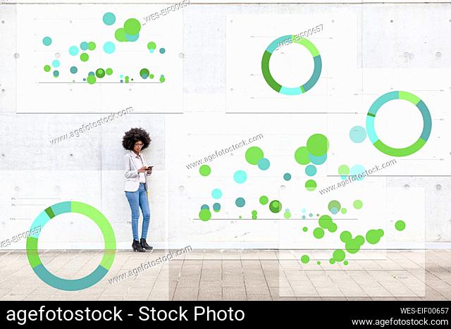 Businesswoman using mobile phone while standing with pie chart icons around on footpath
