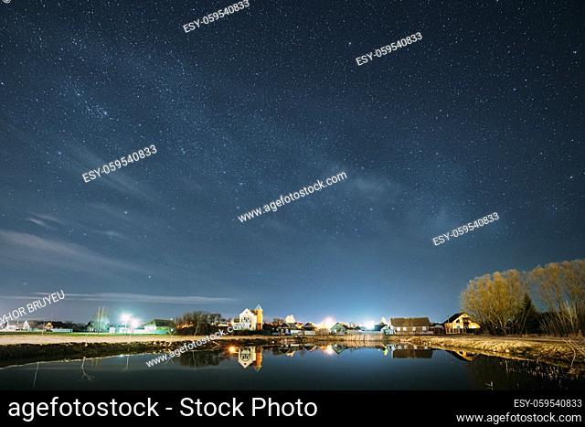 Belarus, Eastern Europe. Night Sky Stars Above Countryside Landscape With Lake Coast And Small Town Or Village. Natural Starry Sky Above Pond And Houses In...