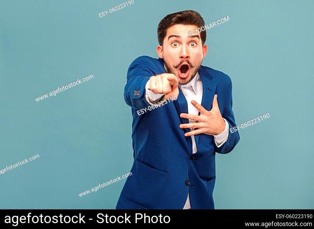 Wondered man pointing finger at camera with shocked face. Business people concept, richly and success. Indoor, studio shot on light blue background