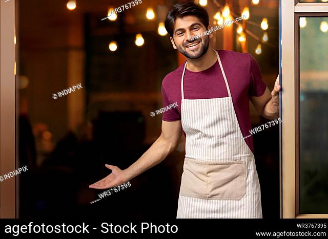 A HAPPY WAITER STANDING IN FRONT OF RESTAURANT AND WELCOMING