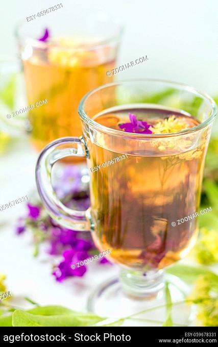 Linden herbal tea in a transparent grog glass with a linden blossom and bunch of herbs on the white wooden surface