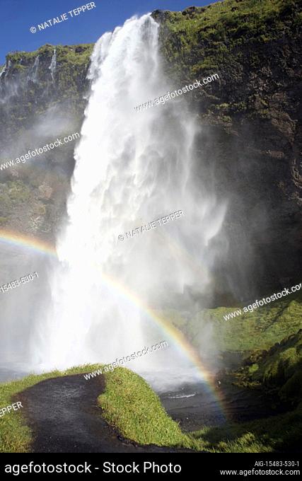 A stunning landscape of a large waterfall called Seljalandsfoss Waterfall , in Iceland, surrounded by fields and mountains, and crowds, and a rainbow