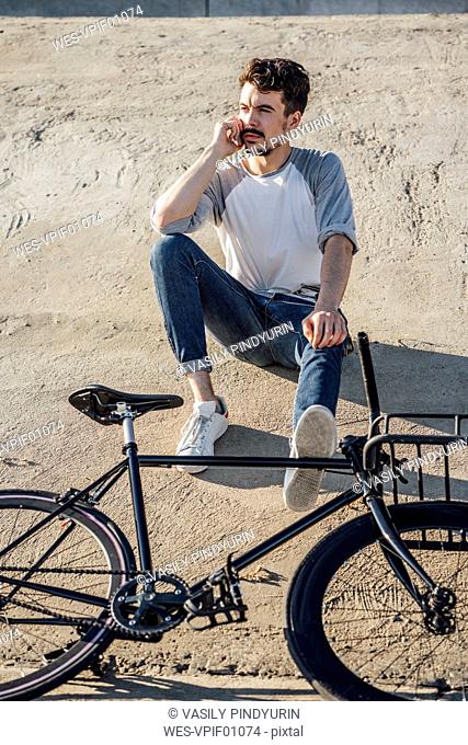Young man with commuter fixie bike sitting on concrete wall talking on cell phone