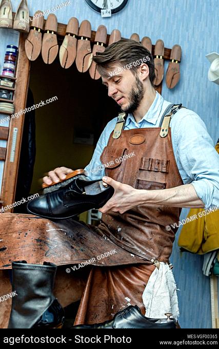 Male cobbler in apron polishing shoe with brush at workshop