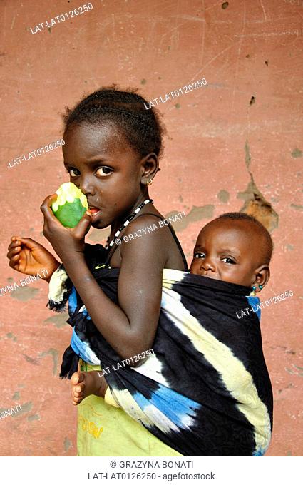 A young girl eating a piece of fruit. Mango. Carrying a baby on her back. In a fabric material sling. Siblings. St Imabe