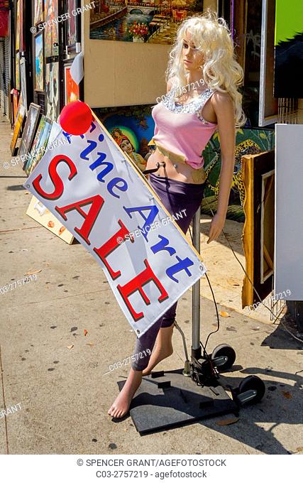 A blonde wigged mannequin displays a sign advertising a fine art sale on the sidewalk outside a Los Angeles store. Note electric motor in mannequin's belly to...