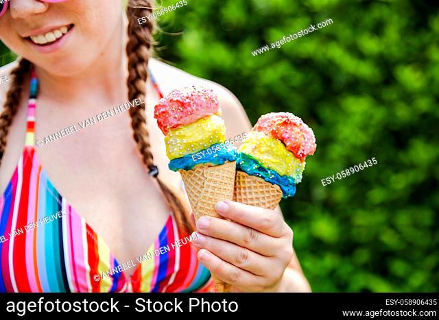 Beautiful girl holding two colorful ice cream with sprinkles wearing a rainbow colored swimsuit in the summer, pink sunglasses and braids happy vacation