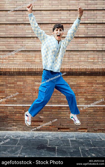 Happy young man jumping on footpath in front of brick wall