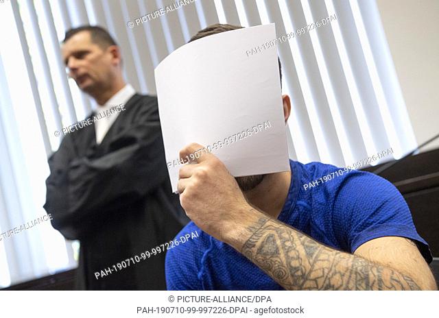 10 July 2019, Hessen, Wiesbaden: When entering the courtroom, the defendant Ali B. covers his face with a piece of paper in front of the journalists' cameras