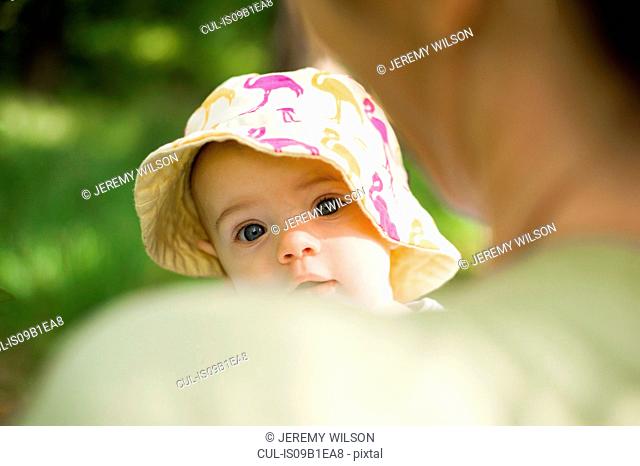Baby girl wearing sunhat looking over mothers shoulder at camera