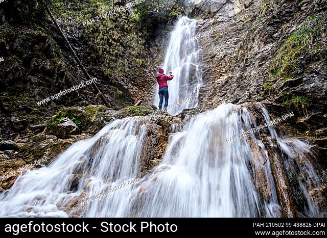 02 May 2021, Bavaria, Grassau: A woman stands at the Grießenbach waterfall in rainy weather and takes a photo with her smartphone