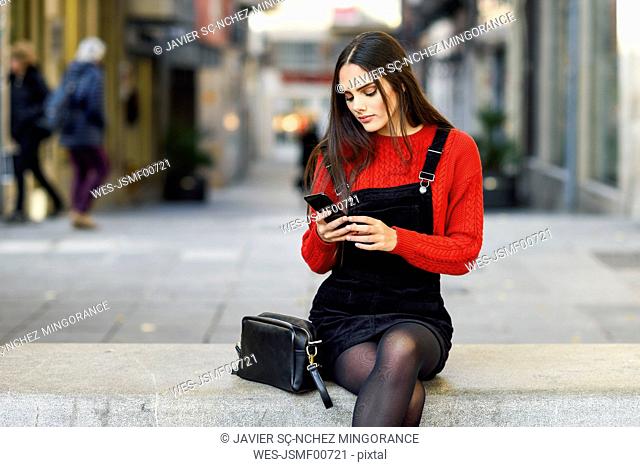 Portrait of fashionable young woman sitting on bench at pedestrian area looking at cell phone