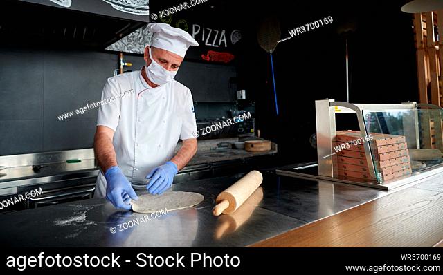 Skilled chef preparing traditional italian pizza in interior of modern restaurant kitchen with special wood-fired oven. Wearing protective medical face mask and...