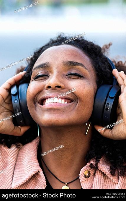 Happy afro woman with eyes closed listening music through headphones in city