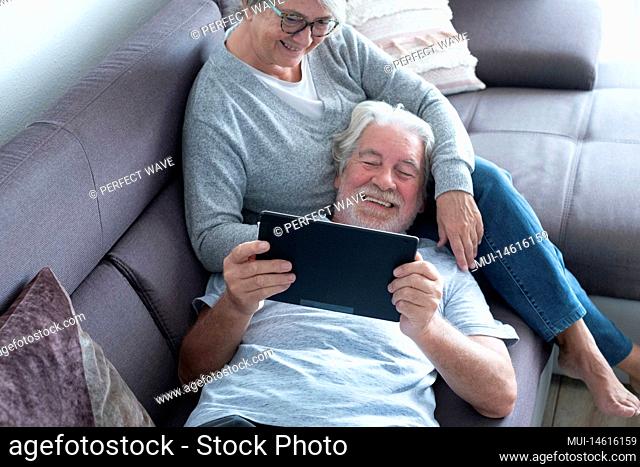 couple of two seniors or mature people at home having fun together looking at the same tablet - pensioner lying on the sofa with his wife sitting