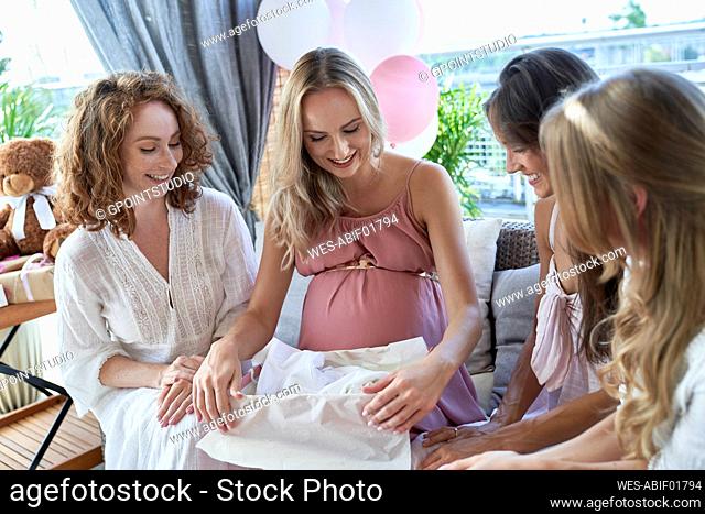 Smiling pregnant woman opening gift box sitting with friends