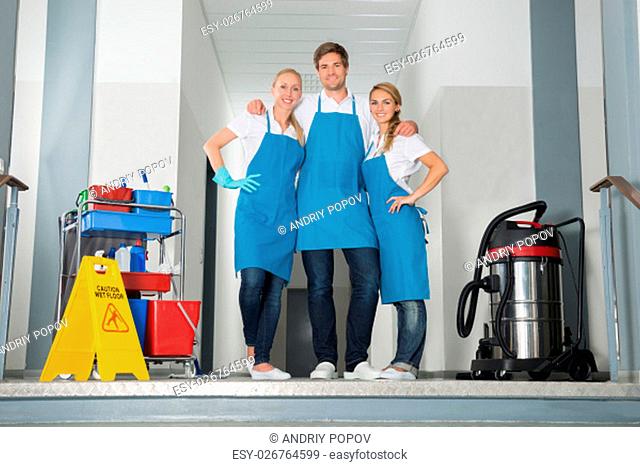 Portrait Of Smiling Janitors Holding Cleaning Equipments By Yellow Wet Caution Sign On Floor