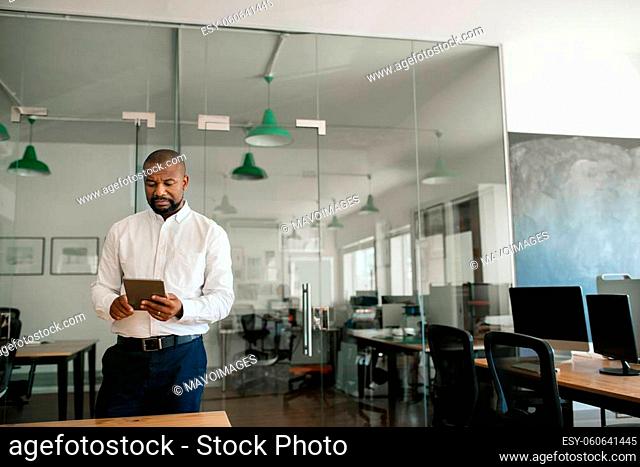 Mature African American businessman using a digital tablet while working alone in a large modern office in the late afternoon
