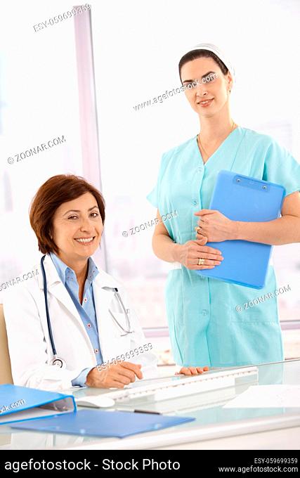 Portrait of senior medical doctor with assistant, smiling at camera