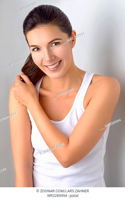 Single cute woman in sleeveless blouse holding ponytails in her hands while standing near gray wall