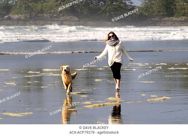 A young woman and her dog (a Golden Retriever) run with a ball on Chesterman Beach near Tofino, BC