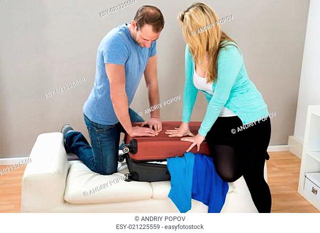 Photo Of Couple Trying To Close Suitcase At Home