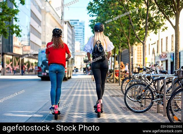 Rear view of trendy fashinable teenager girls riding public rental electric scooters in urban city environment. New eco-friendly modern public city transport in...