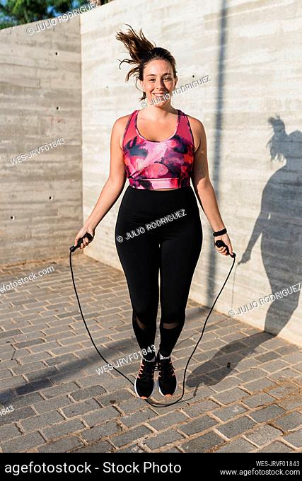 Sportswoman smiling while skipping by wall during sunny day