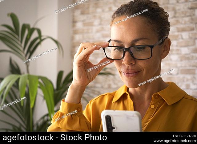 Confident portrait of young pretty adult woman smiling and using mobile phone connection at home or office. Attractive female people wearing eyeglasses and...