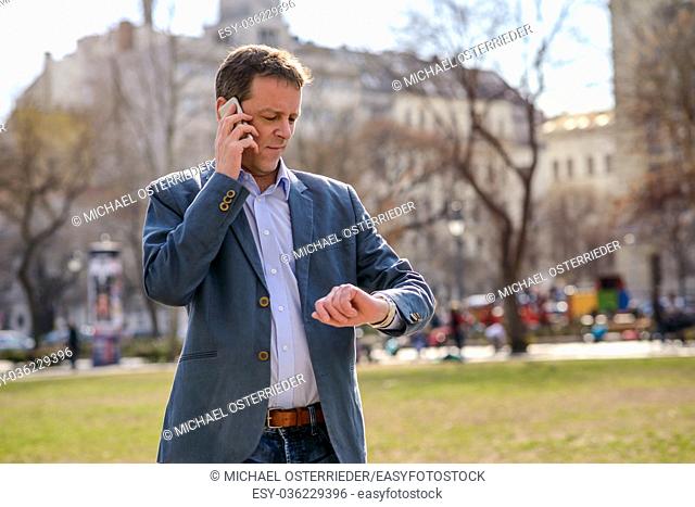 A middle age businessman standing in a park while talking on his phone and checking the time on his watch