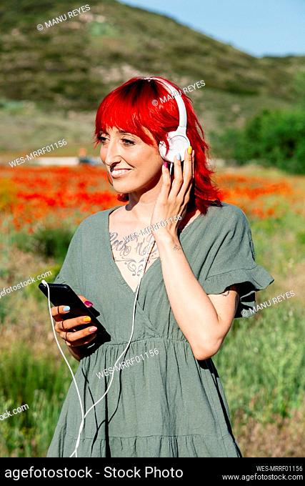 Smiling young woman with mobile phone listening music through headphones on poppy field