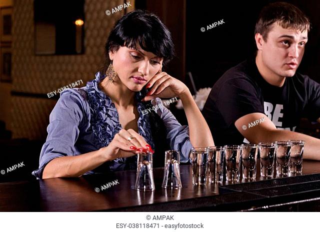 Lonely woman alcoholic sitting at a bar counter with a long line of full shot glasses staring morosely at the counter as she drinks her way through them
