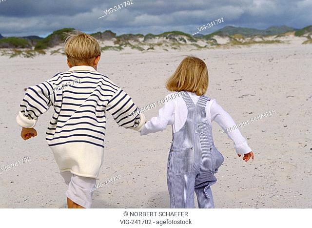 beach-scene, couple viewed from the back, blond girl wearing trousers with braces and blond boy with striped pullover run hand in hand bare feeted through the...