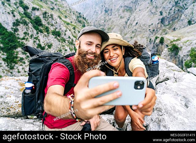 Couple smiling while taking selfie sitting on mountain peak at Ruta Del Cares, Asturias, Spain