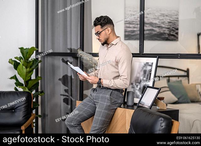 Study of documents. Young bearded man in glasses standing sideways to camera reading examining documents at home