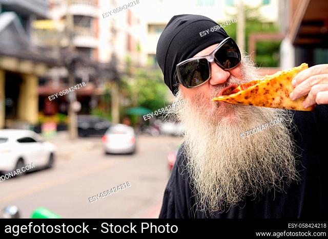 Portrait of mature bearded man eating slice of New York style pepperoni pizza outdoors