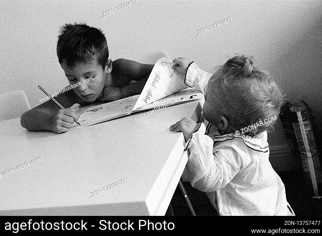 A boy sitting at the table and doing his homework. His curious baby sister is standing nearby and holding a page of his book