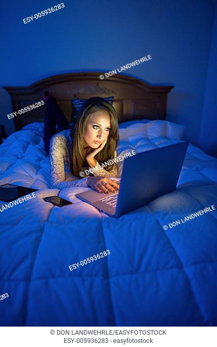 Teenage girl lying on a bed at night working on a laptop computer