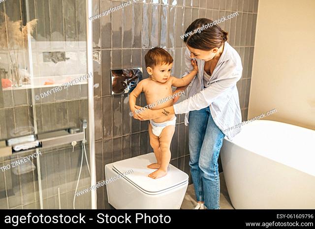 Caring female parent supporting her child in the diapers standing on the closed WC lid