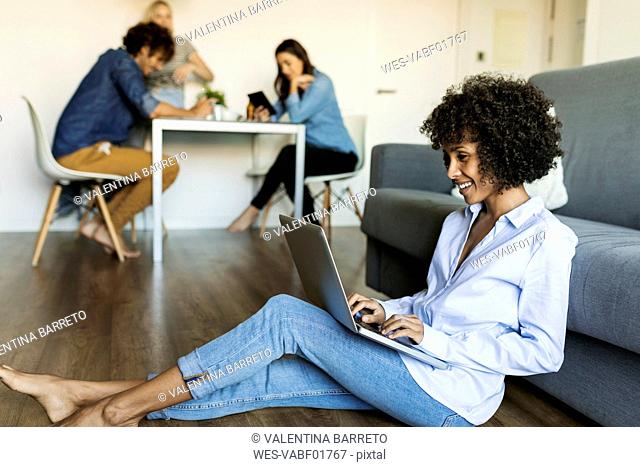 Smiling woman sitting on floor using laptop with friends in background