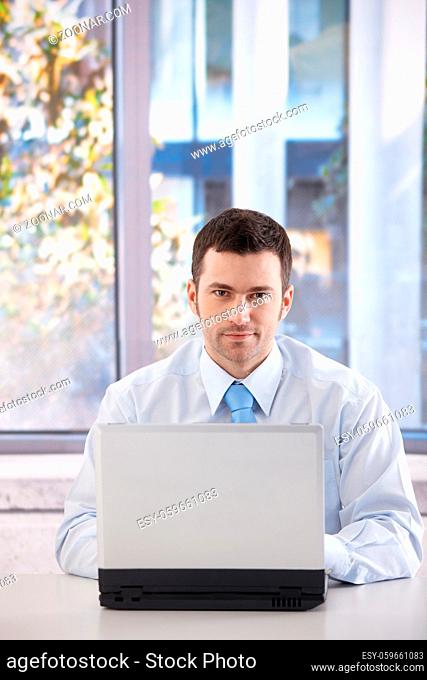 Portrait of young confident businessman working on laptop in bright office