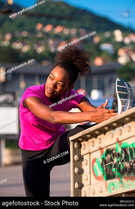 young beautiful African American woman doing warming up and stretching with her leg raised to the bridge before the morning run with the sunrise in the...