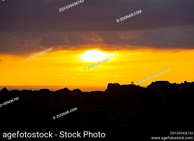 Silhouette of two people standing on distant ridge watching sun breaking thru clouds making a colorful orange sky at sunset in Gobi Desert in rural Mongolia