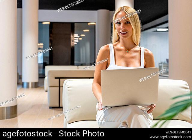 Smiling blond businesswoman with laptop sitting on sofa in office