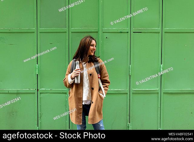 Smiling young woman holding thermos flask in front of a green wall