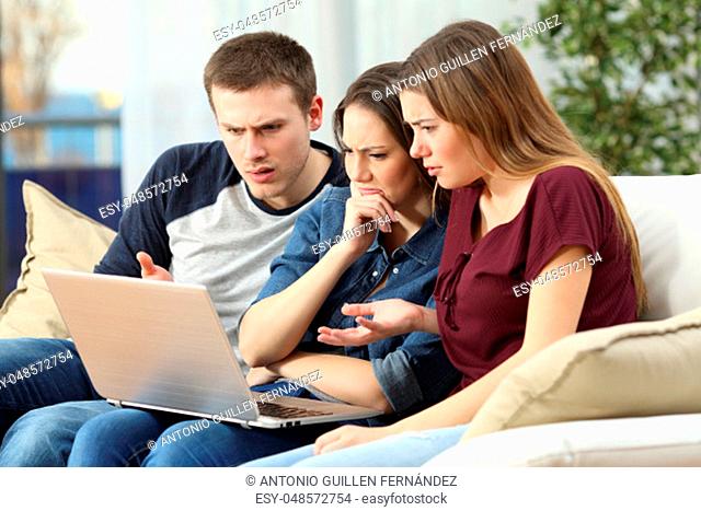Three angry friends watching media content on line in a laptop sitting on a couch in the living room in a house interior