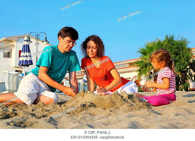 Mother with father and girl sit in day-time on beach and build hills from sand