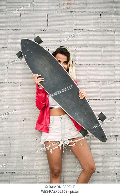 Portrait of a young woman with her long board, in front of a wall