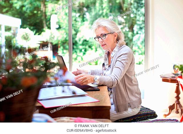 Senior woman sitting at table, looking through paperwork, using laptop